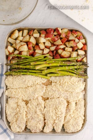 breaded chicken and asparagus arranged on a baking sheet with par roasted potatoes