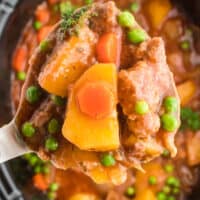 close up of beef stew in a crock pot with a spoon full of the soup on a big metallic spoon