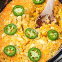 square image of a wooden spon in slow cooker cheddar jalapeno corn casserole with jalapeno slices on top