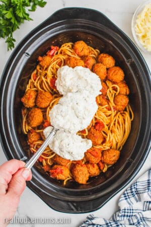 hand suing a spoon to add dollops of the ricotta mixture to the crockpot of spaghetti and meatballs