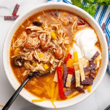 square image of a spoon in a bowl of slow cooker chicken taco soup topped with sour cream, tortilla strips, and cheddar cheese