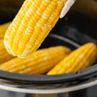 tongs lifting an ear of slow cooker corn on the cob from the crock pot with recipe name at the bottom