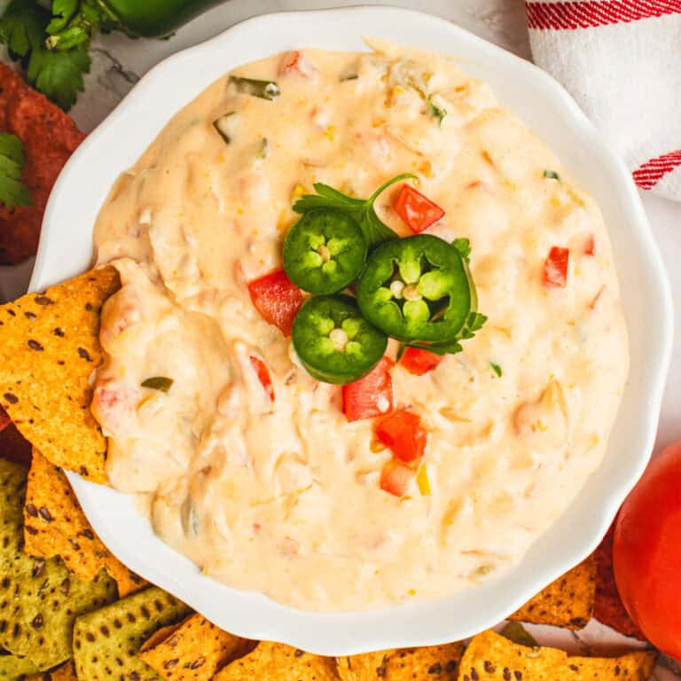 square image of slow cooker queso dip topped with jalapeno slices and diced tomatoes in a serving bowl