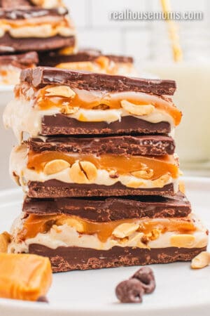 stack of snickers cheesecake bars on a white plate next to chocolate chips and caramels