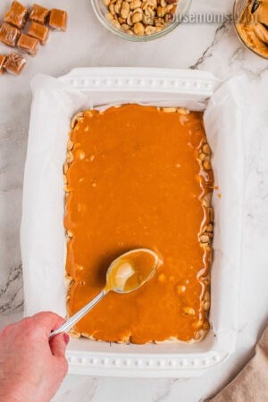 melted caramel being spread over peanut layer for snickers cheesecake bars