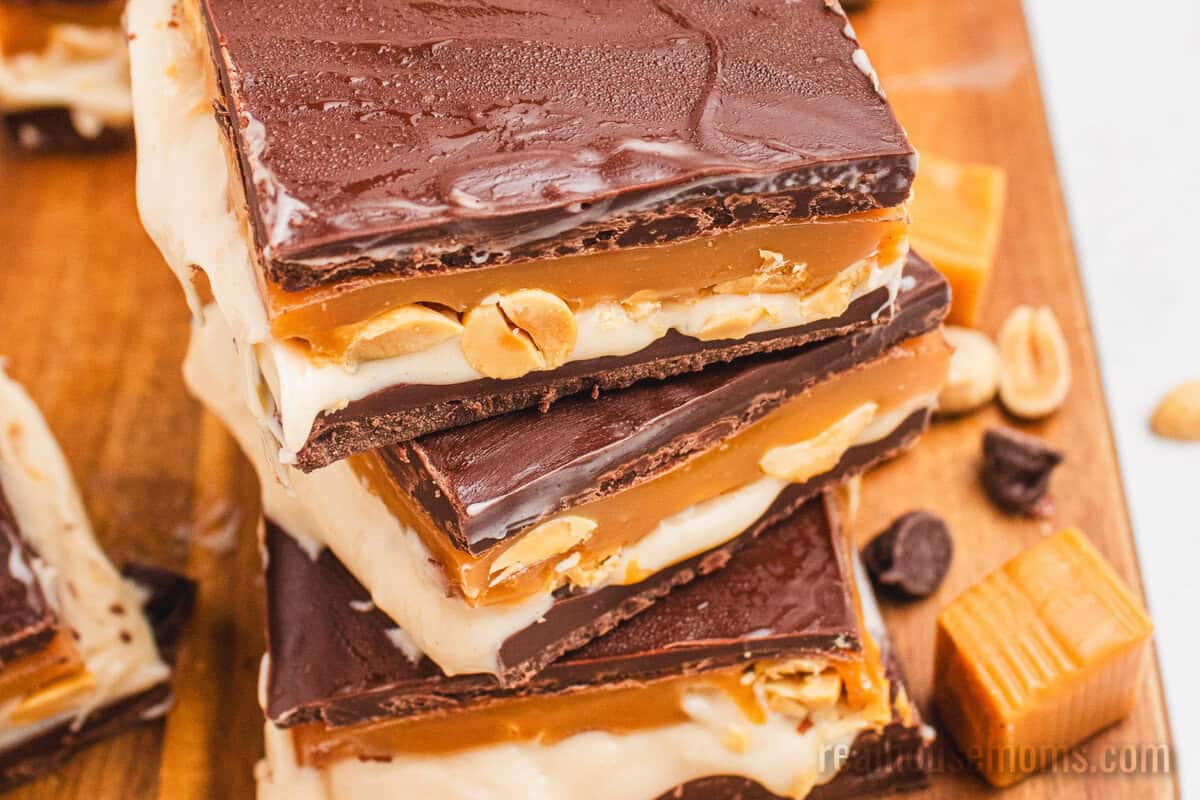 angled view of snickers cheesecake bars stacked on a wooden cutting board