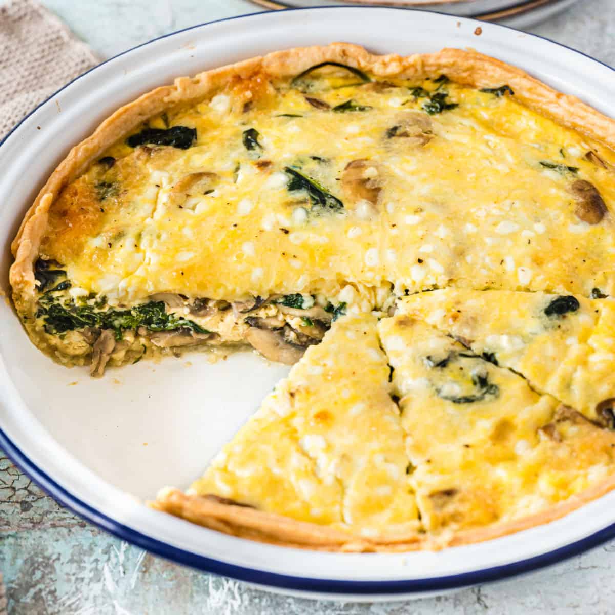 square image of a spinach and mushroom quiche with a slice taken out of the plate to show the filling