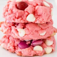 stack of strawberry cake mix cookies in front of a plate of cookies with a bottle of milk with recipe name at bottom