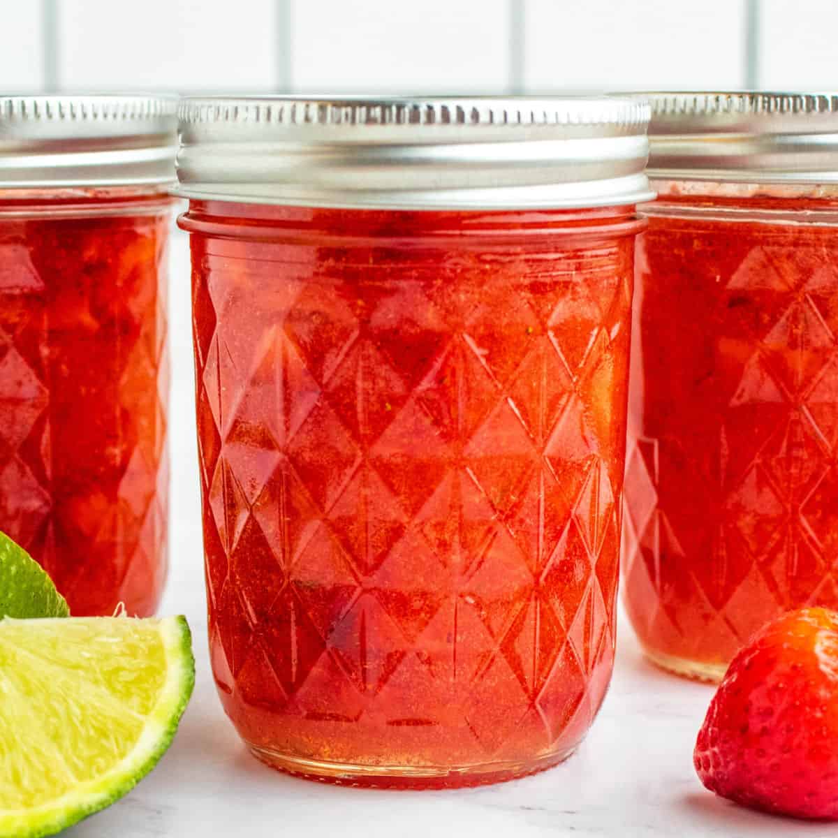 square image of jars of strawberry freezer jam next to limes and strawberries