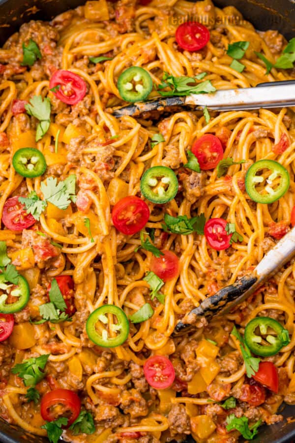 skillet of taco spaghetti topped with tomatoes, jalapeno, and cilantro with tongs for serving