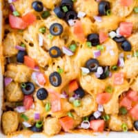 square close up image of taco tater tot casserole topped with tomatoes, red onion, and olives
