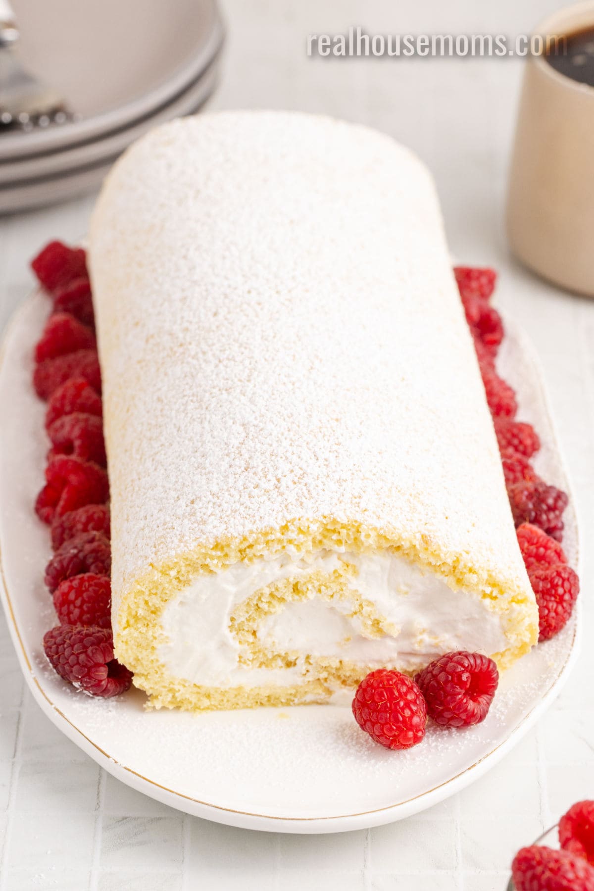 vanilla swiss roll coated in powdered sugar on a platter and surrounded by raspberries