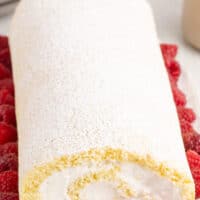 image vanilla swiss roll coated in powdered sugar on a platter and surrounded by raspberries with the title of the post on top of the image in pink and black lettering