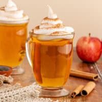 square image of mugs of warm apple pie cocktail topped with whipped cream and cinnamon next to an apple