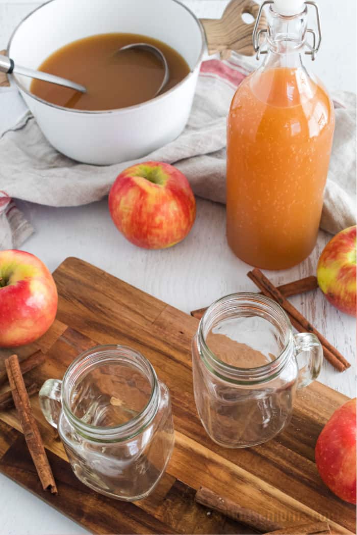 boozy apple cider in a pot with a ladle, next to a carafe of n/a apple cider and empty mugs