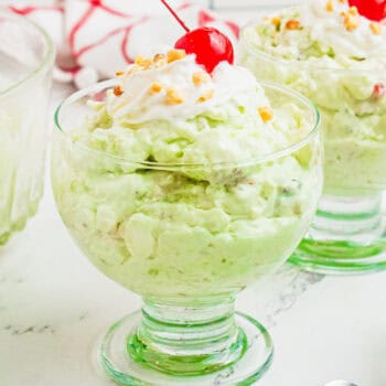 square image of watergate salad in a dessert dish topped with whipped cream, chopped nuts, and a cherry