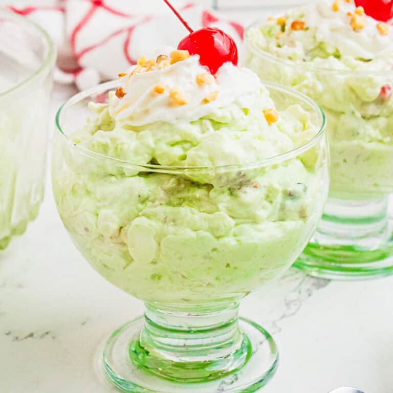 square image of watergate salad in a dessert dish topped with whipped cream, chopped nuts, and a cherry