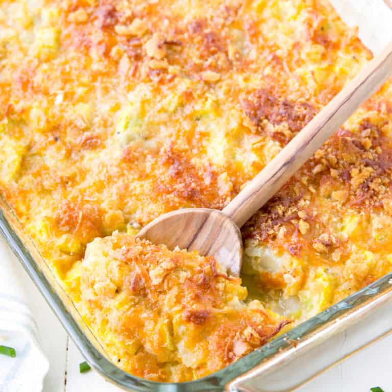 square image of yellow squash casserole in a glass baking dish with a wooden spoon