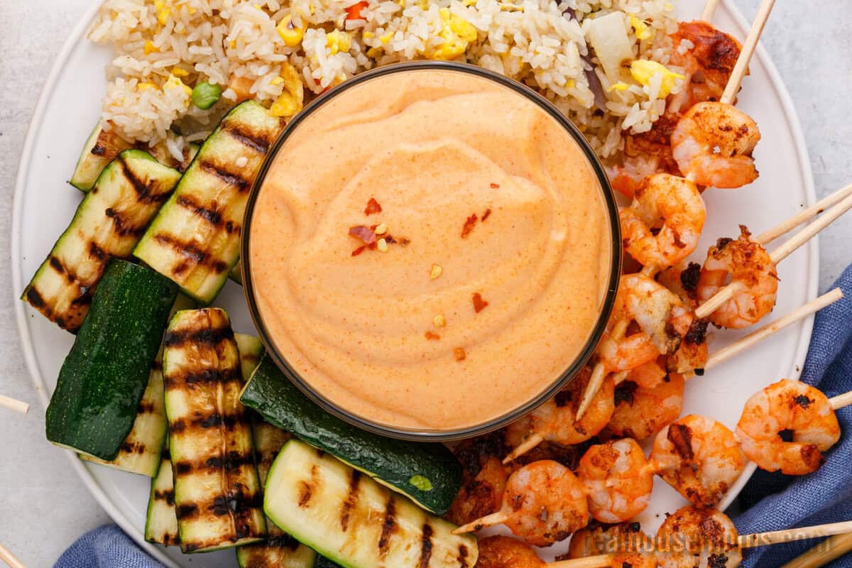looking down at a bowl of yum yum sauce on a plate with fried rice, grilled zucchini, and shrimp skewers